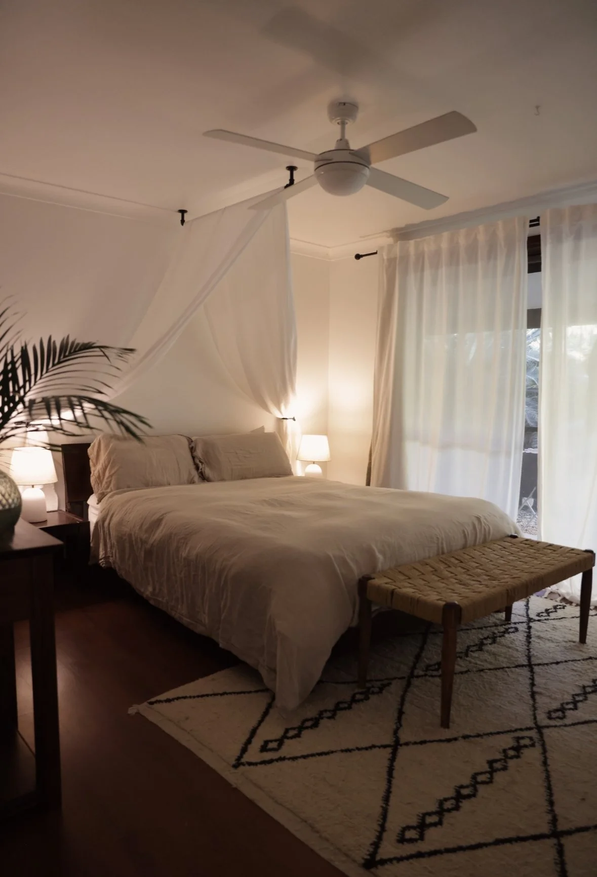 Bedroom after — canopy detail, warm lamplight, Moroccan rug, rattan bench, layered soft textiles